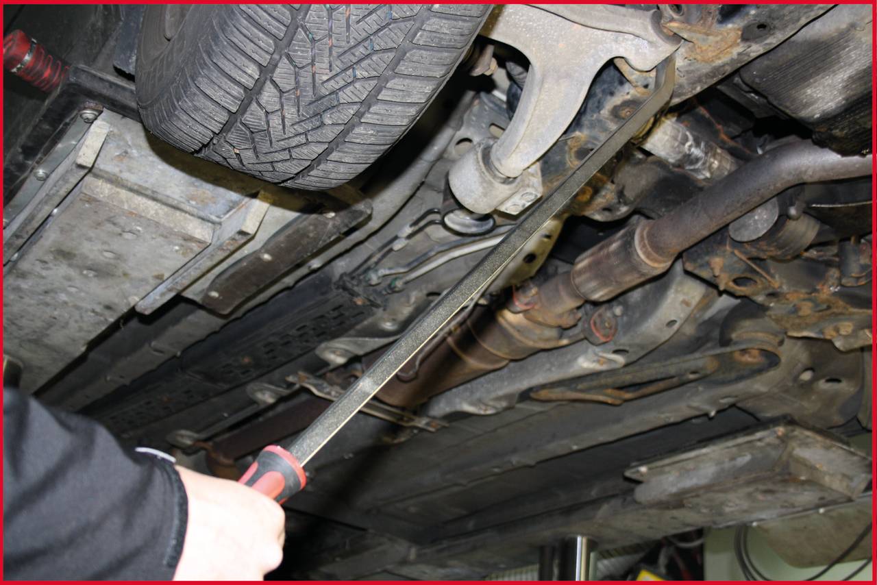 Ansicht einer Autounterseite in einer Werkstatt. Ein Mechaniker nutzt ein Werkzeug, um einen Bereich zu kontrollieren. Reifen und Abgasanlage sichtbar.