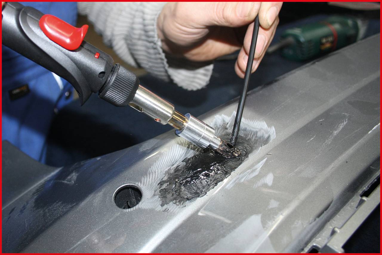 Person repariert ein Metallteil mit einer Heißluftpistole und Füllmaterial. Fokus liegt auf präziser Arbeit, um Schaden zu beheben.
