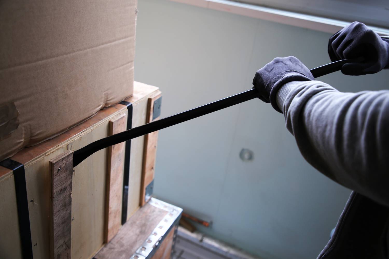 Eine Person mit Handschuhen öffnet mit einem Brecheisen eine Holzkiste, die auf einem Kartonstapel im Innenraum steht.