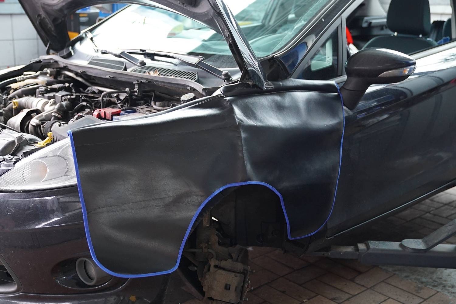 Ein Auto in einer Werkstatt, die Motorhaube offen, mit einer Schutzabdeckung über einem Kotflügel, sichtbar sind Motor und Werkzeuge.