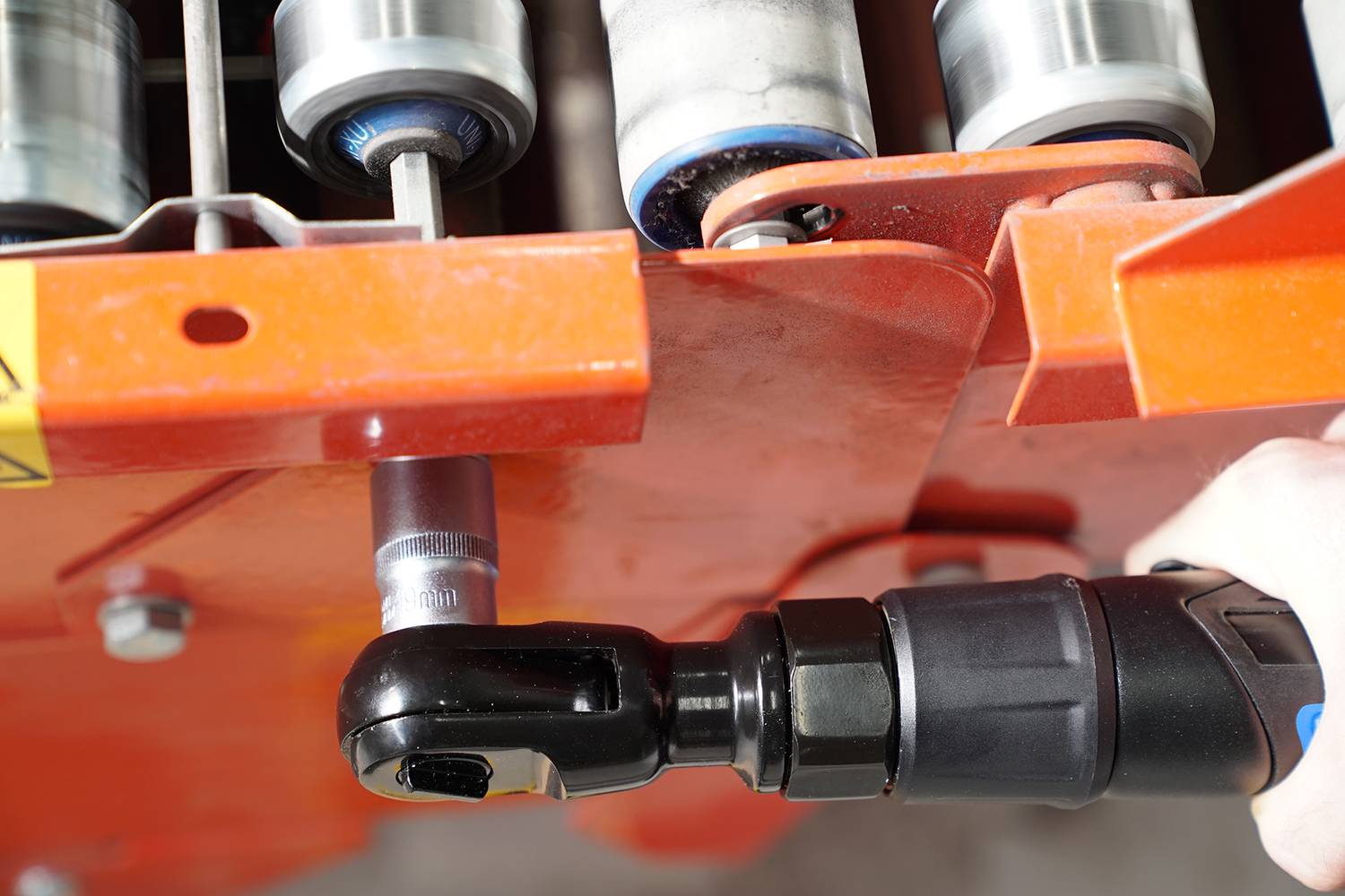 Nahaufnahme eines orangenen Maschinenbauteils, das mit einem schwarzen Werkzeug bearbeitet wird. Im Hintergrund sind Rollen sichtbar.