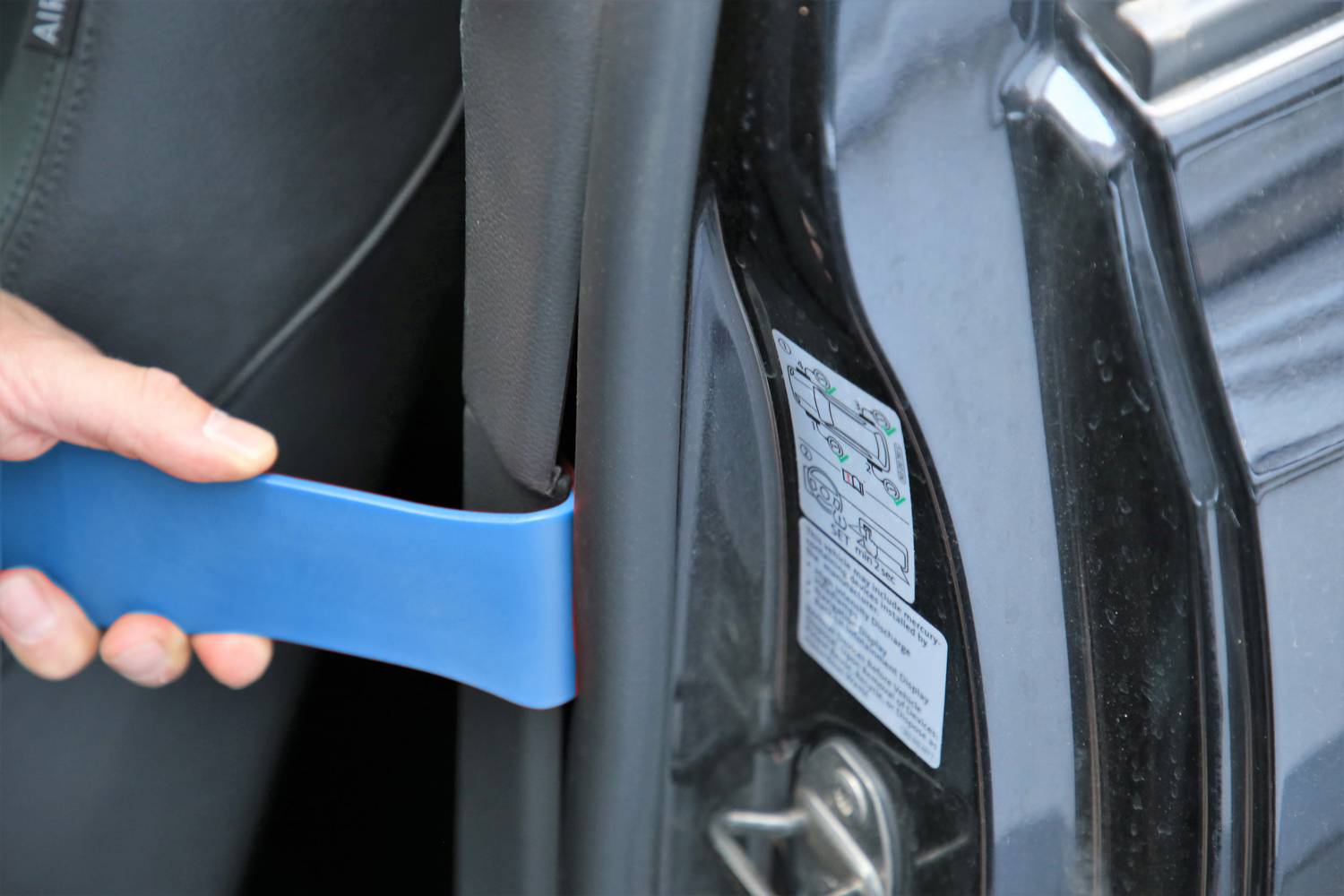 Eine Person verwendet ein blaues Kunststoffwerkzeug, um die Autotürverkleidung zu entfernen. Ein Hinweisschild ist an der Tür angebracht.