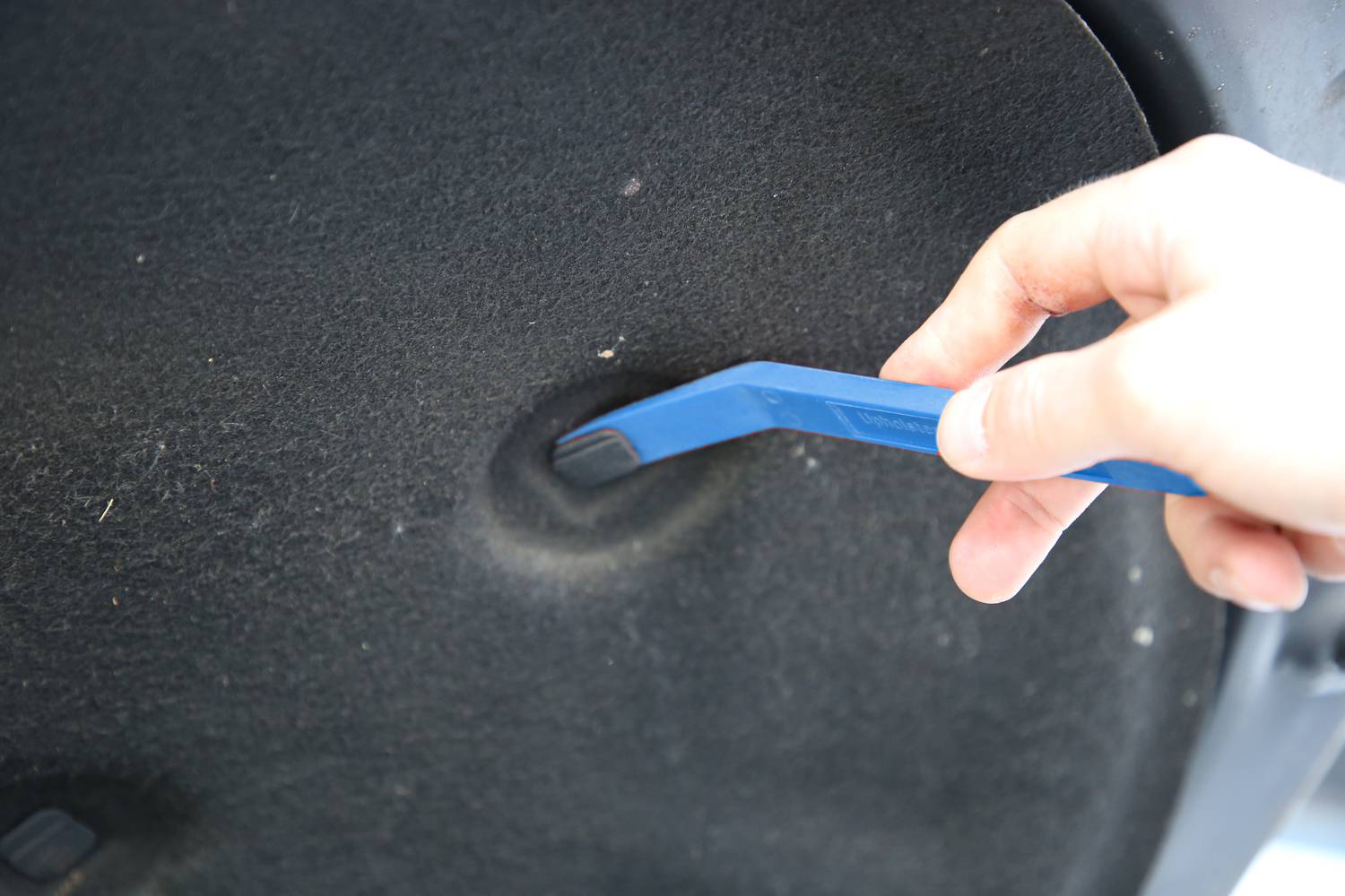 Eine Hand hält ein blaues Kunststoffwerkzeug und entfernt vorsichtig eine Abdeckung in einem Auto, vermutlich zur Innenreparatur.