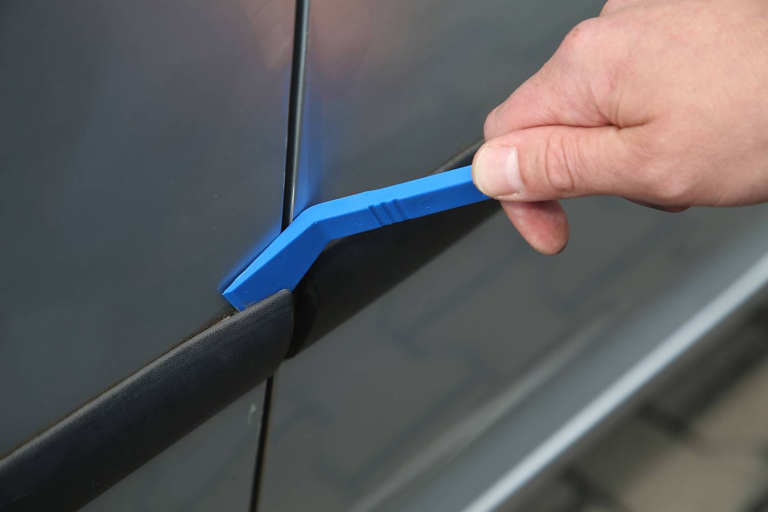 Eine Hand verwendet ein blaues Kunststoffwerkzeug, um den Spalt zwischen einem Autotürrahmen und einer Dichtung zu öffnen.