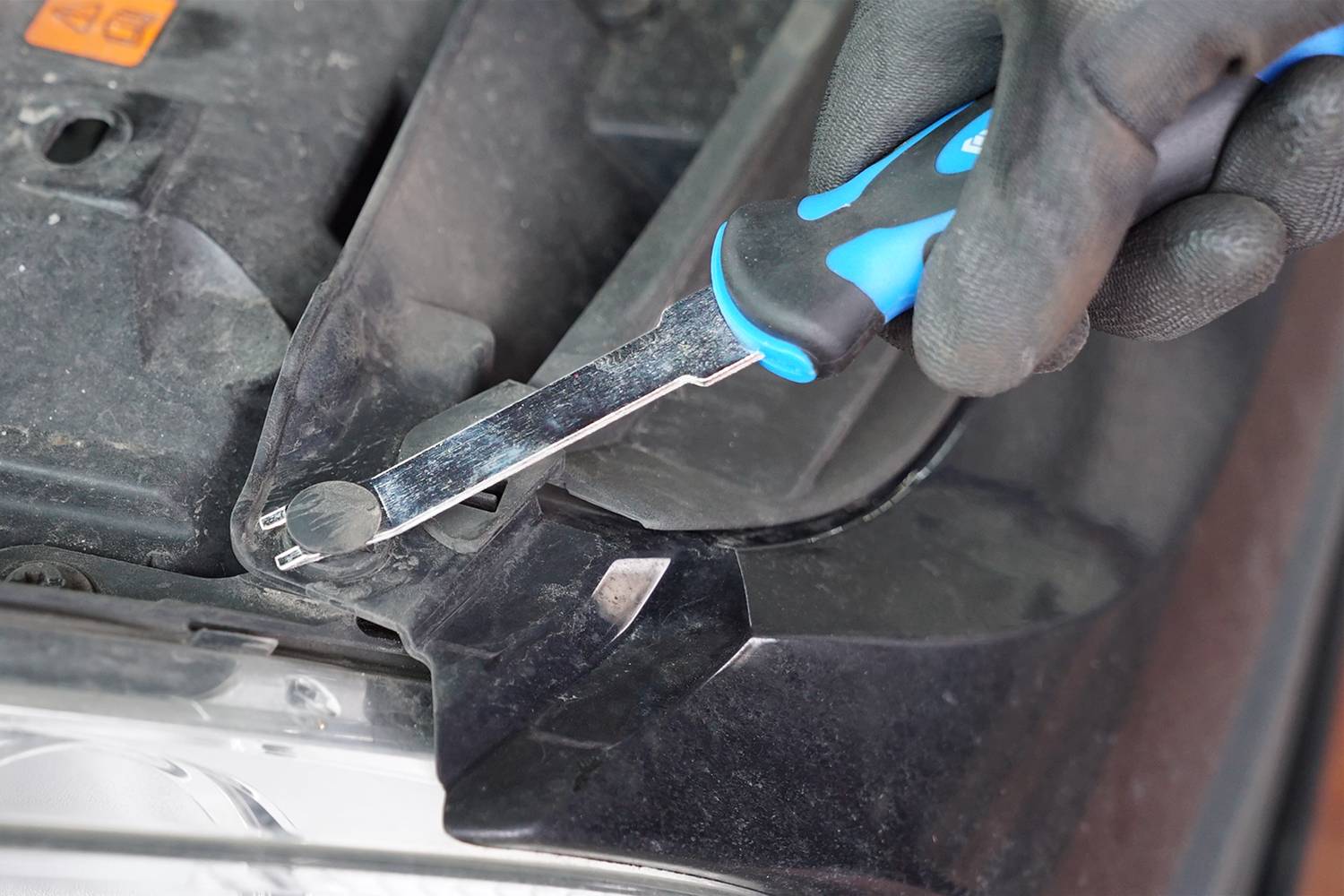 'Nahaufnahme einer Hand mit Arbeitshandschuhen, die ein Spezialwerkzeug verwendet, um eine Kunststoffverkleidung im Vorderbereich eines Autos zu entfernen.'