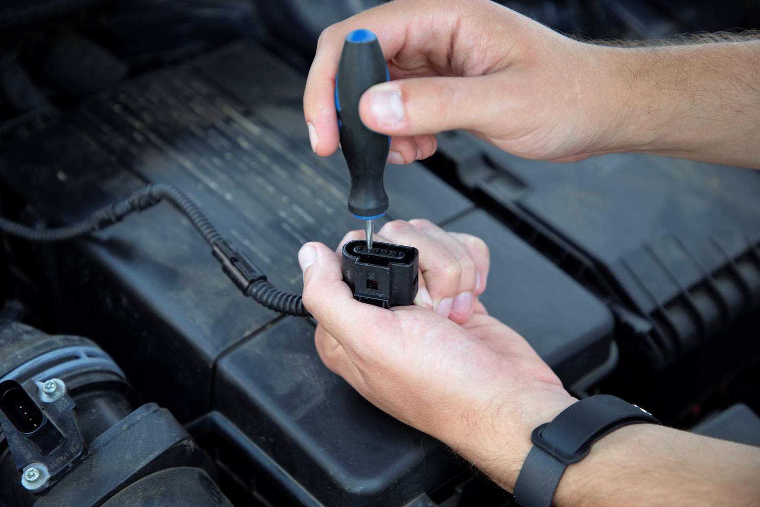 Person repariert einen elektrischen Stecker im Motorraum eines Autos mit einem Schraubenzieher. Hände fokussiert, Hintergrund unscharf.