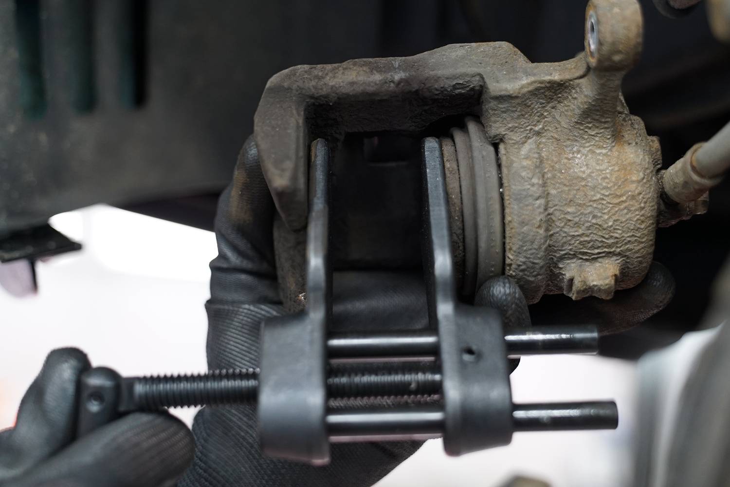 Eine Hand mit Handschuh benutzt ein Werkzeug, um einen Bremssattel zurückzudrücken. Deutliche Werkstattumgebung erkennbar.