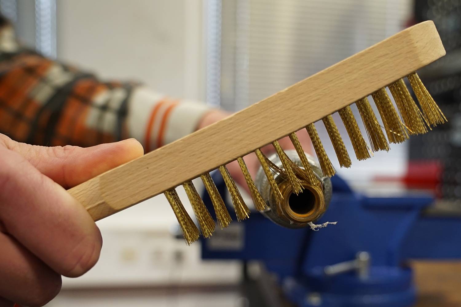 Eine Hand hält eine Drahtbürste neben ein rundes Objekt in einer Werkstatt. Im Hintergrund ist eine Werkbank zu sehen.