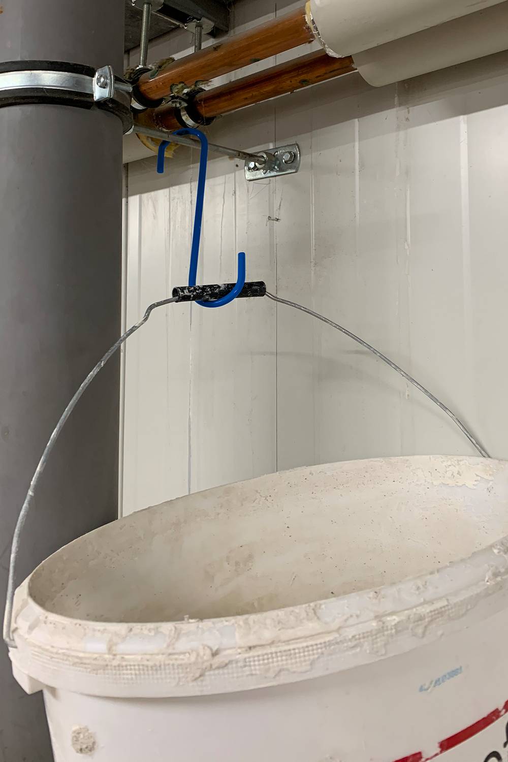 Ein weißer Eimer hängt an einer blauen Hakenkonstruktion, die an einer Rohrleitung befestigt ist. Der Eimer sammelt offenbar tropfendes Wasser.