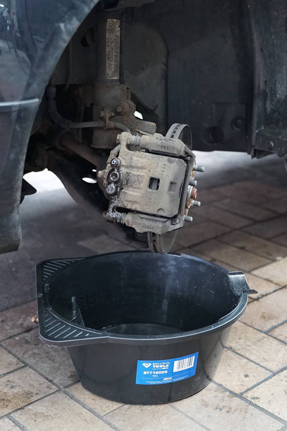 Ein Auto steht aufgebockt, mit demontiertem Reifen und freigelegter Bremse. Darunter steht ein schwarzer Eimer auf dem Boden.