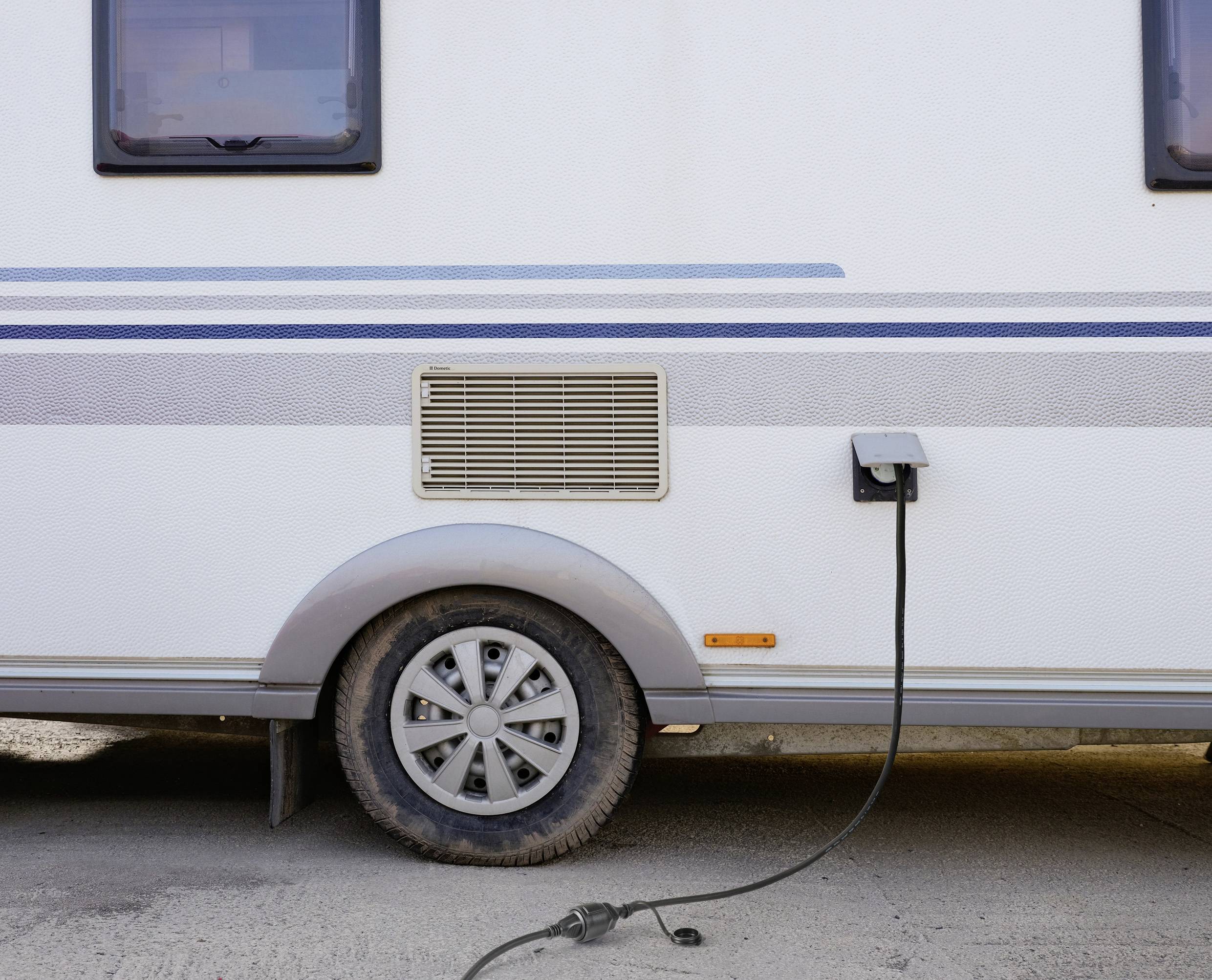 Ein weißer Wohnwagen ist über ein Kabel mit einer Stromquelle verbunden. Das rechte hintere Rad ist teilweise im Bild sichtbar.