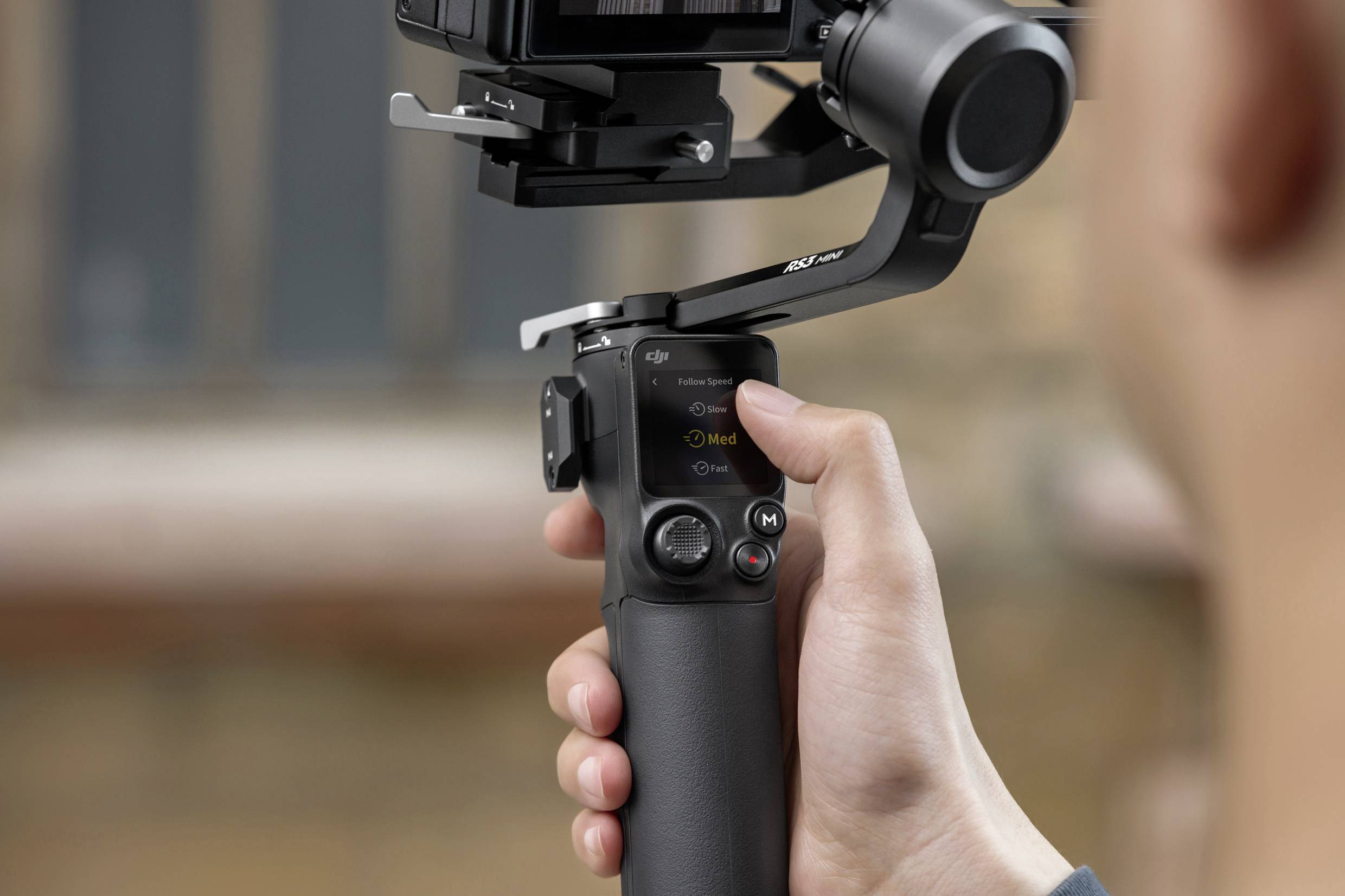 Eine Person hält einen Kameragimbal mit einem kleinen Display, das Einstellungen zeigt. Der Gimbal stabilisiert eine Kamera für Videoaufnahmen.
