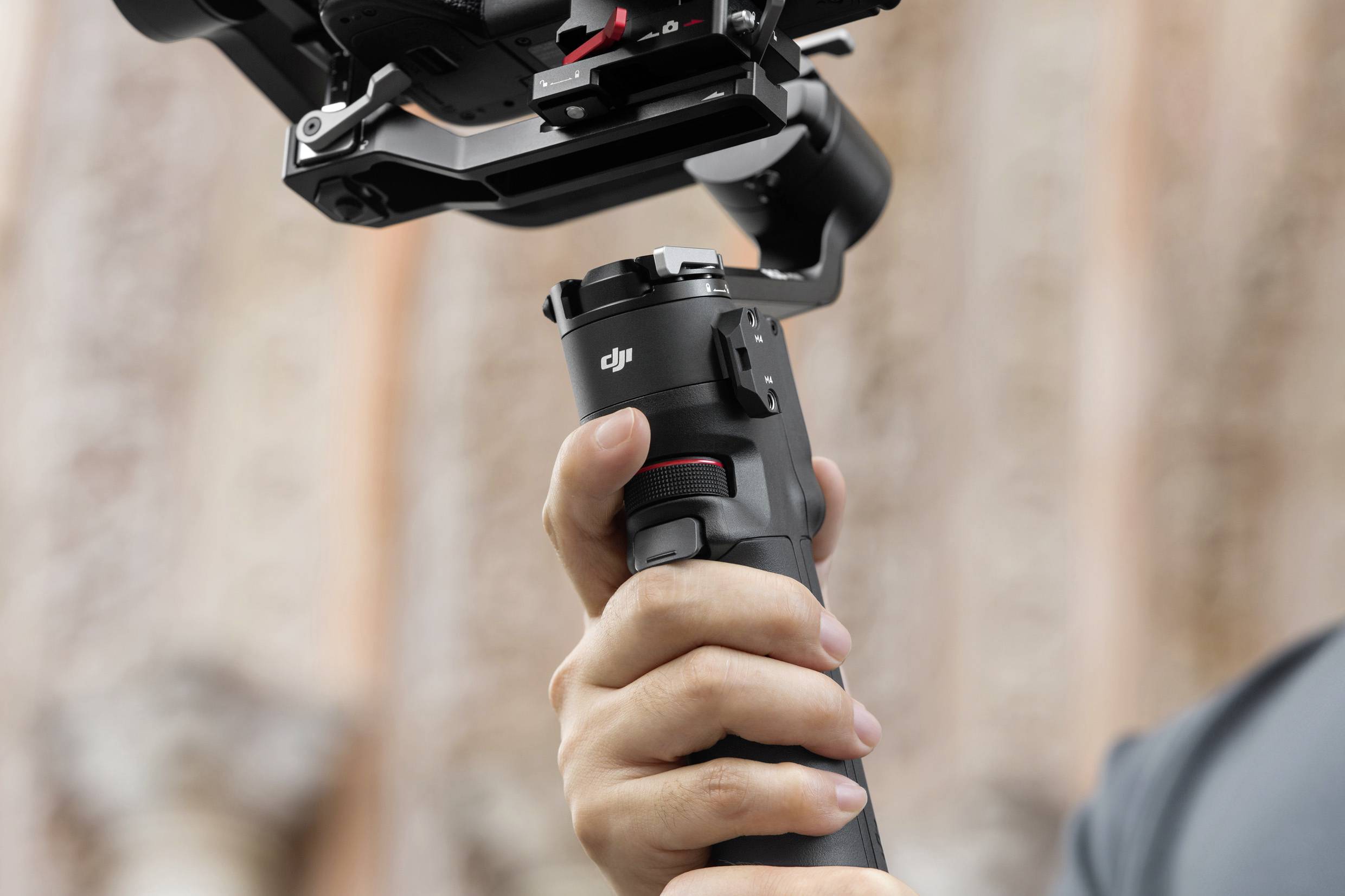 Eine Hand hält den Griff eines professionellen Kamera-Gimbals, im Hintergrund ist eine verschwommene architektonische Struktur zu sehen.