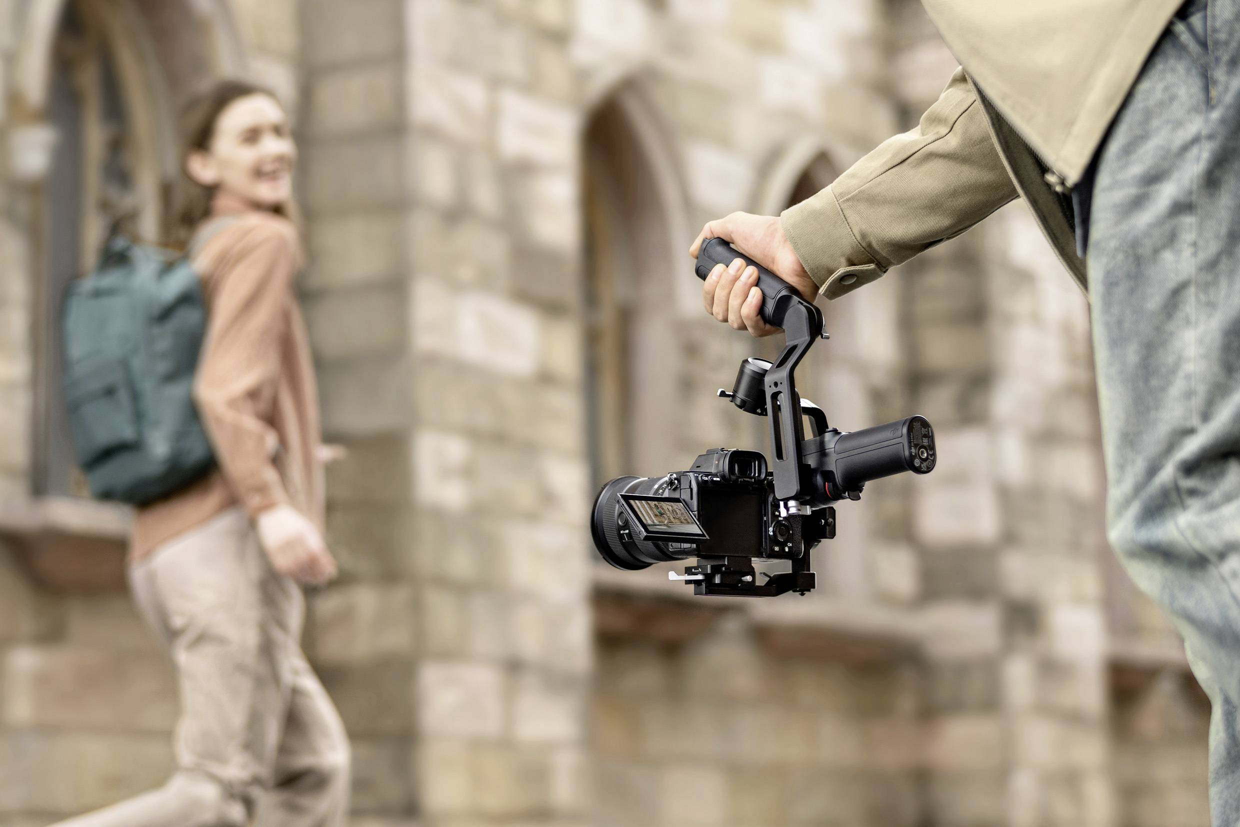 Eine Person filmt mit einer Kamera auf einem Stabilisator, während eine andere Person im Hintergrund vorbeigeht. Hintergrund aus Steinmauer.