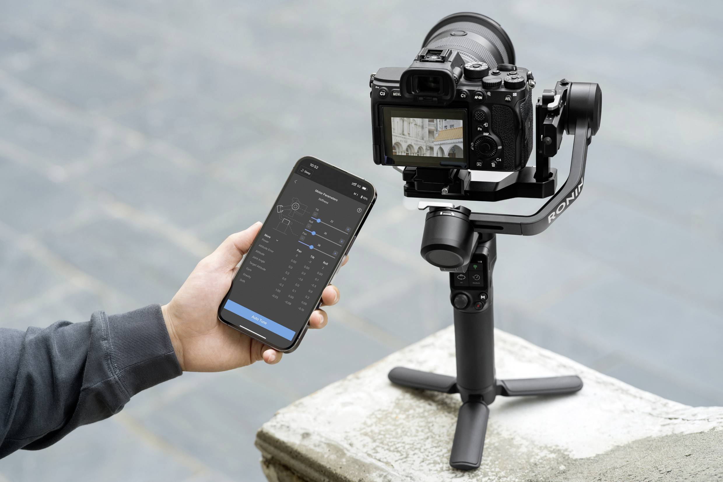 Eine Person hält ein Smartphone mit einer App, die eine Kamera auf einem Gimbal steuert. Die Kamera ist auf einen Platz ausgerichtet.