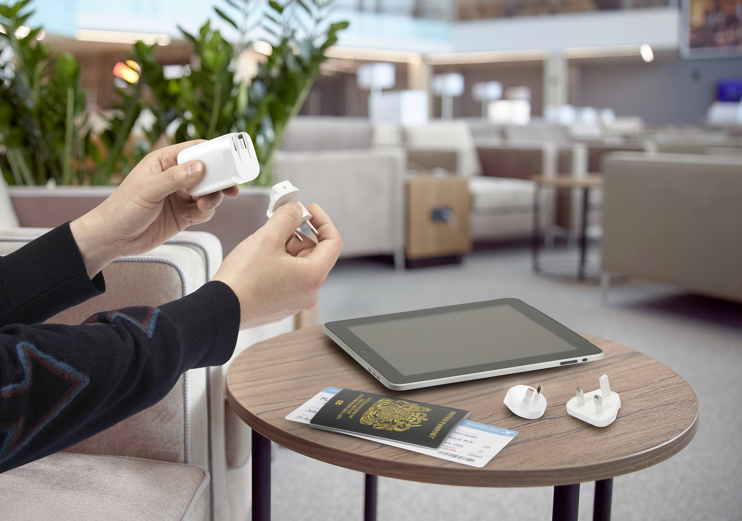 Person hält einen Reiseadapter in einem Wartebereich; auf dem Tisch liegt ein Tablet, ein Pass und ein Boardingpass.