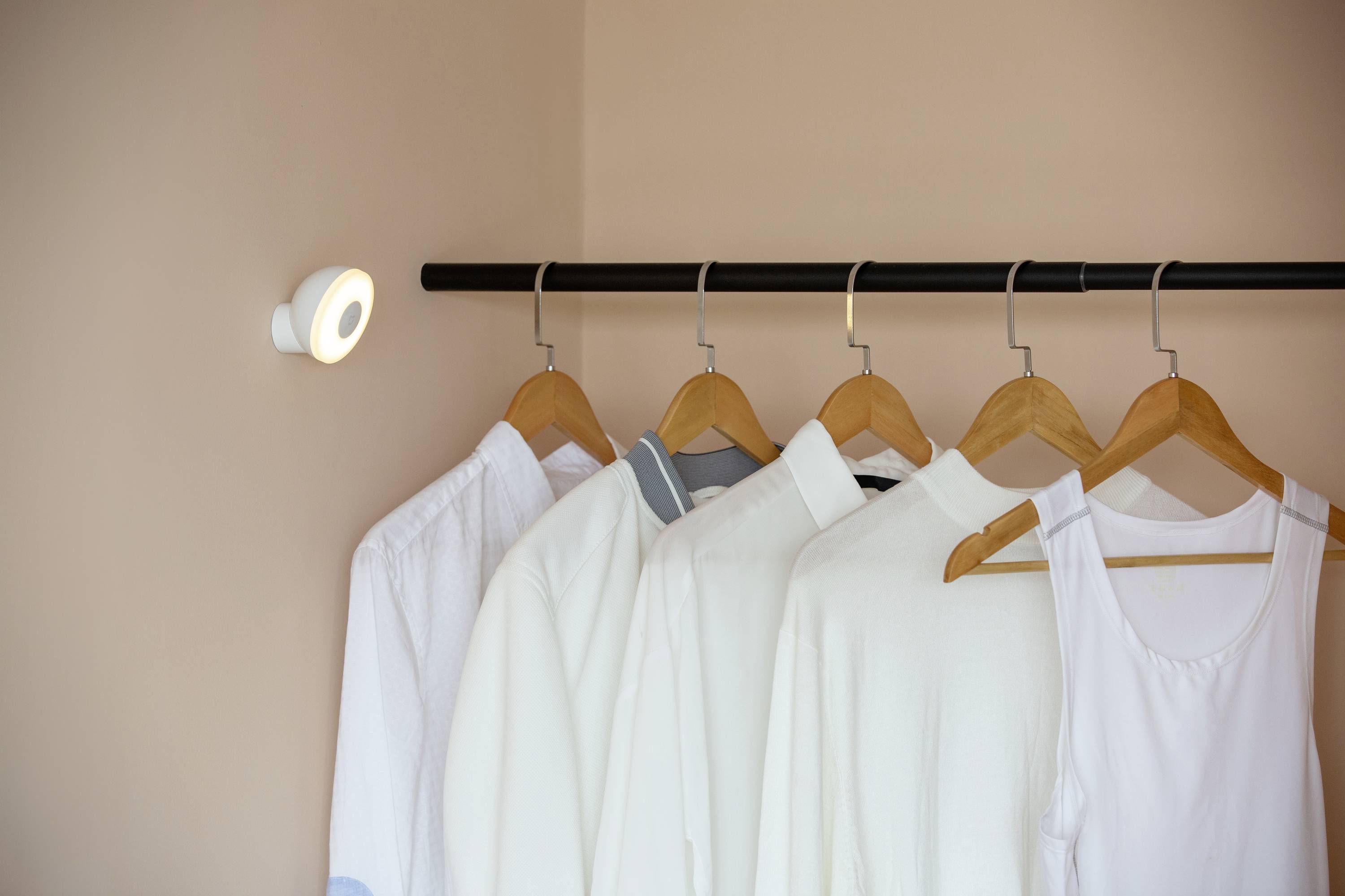 Fünf helle Hemden und ein Tanktop hängen auf Holzkleiderbügeln an einer schwarzen Stange. Links ist eine kleine, leuchtende Lampe.