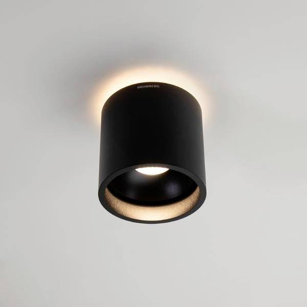 Eine schwarze Deckenleuchte mit zylindrischer Form, die auf einer weißen Decke montiert ist und warmes Licht nach unten abstrahlt.