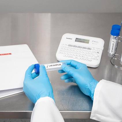 Person in blauen Handschuhen etikettiert ein Röhrchen mit 'Lab Samples' an einem Tisch mit einem Etikettendrucker und Ordnern.