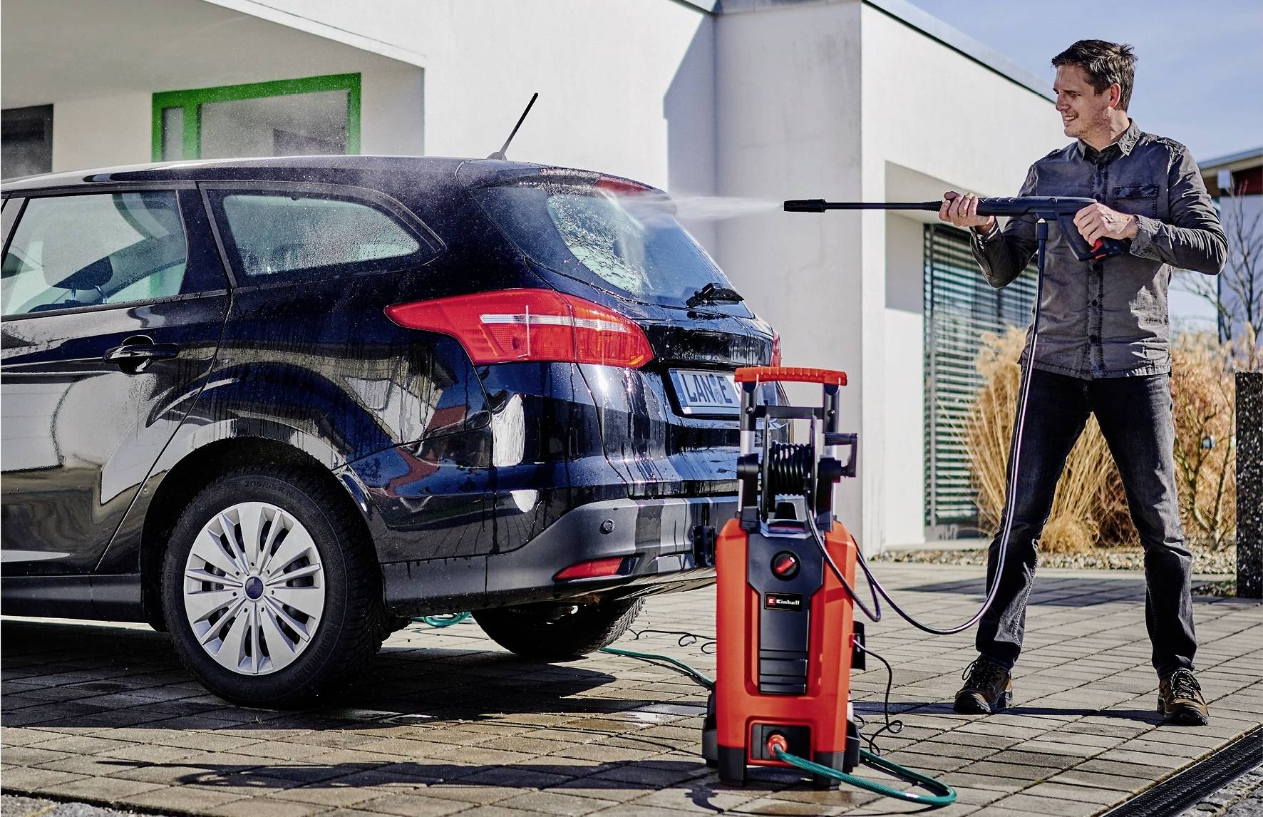 Ein Mann benutzt einen Hochdruckreiniger, um ein schwarzes Auto im Freien zu waschen. Der Hochdruckreiniger ist rot und steht auf dem Boden.