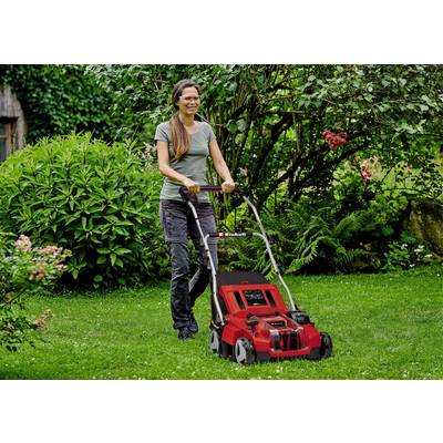 Eine Frau mäht den Rasen mit einem roten Rasenmäher in einem grünen Garten. Sie trägt eine graue Hose und ein grünes T-Shirt.