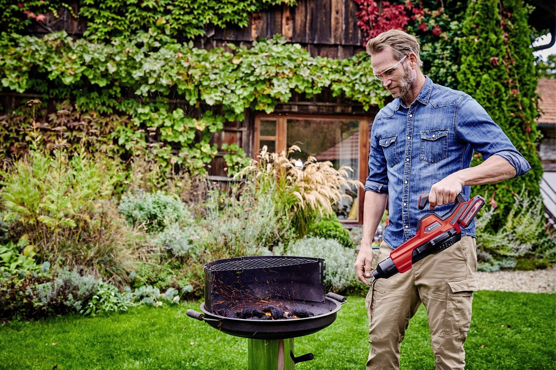 Ein Mann im Garten zündet mit einem elektrischen Grillanzünder den Grill an. Im Hintergrund sind Pflanzen und ein Holzhaus zu sehen.