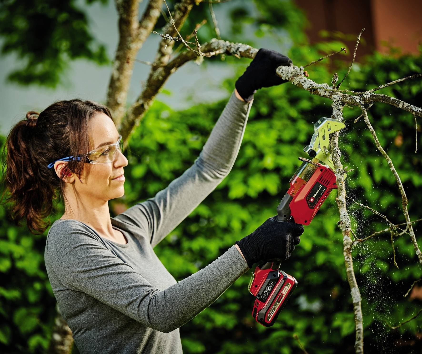 Eine Person mit Schutzbrille und Handschuhen schneidet mit einer elektrischen Astsäge Äste von einem Baum ab.