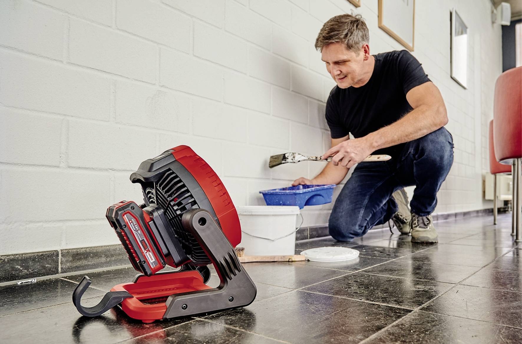 Mann in schwarzem T-Shirt und Jeans streicht eine Wand weiß. Im Vordergrund steht ein kleines, tragbares Lüftungsgerät auf dem Boden.