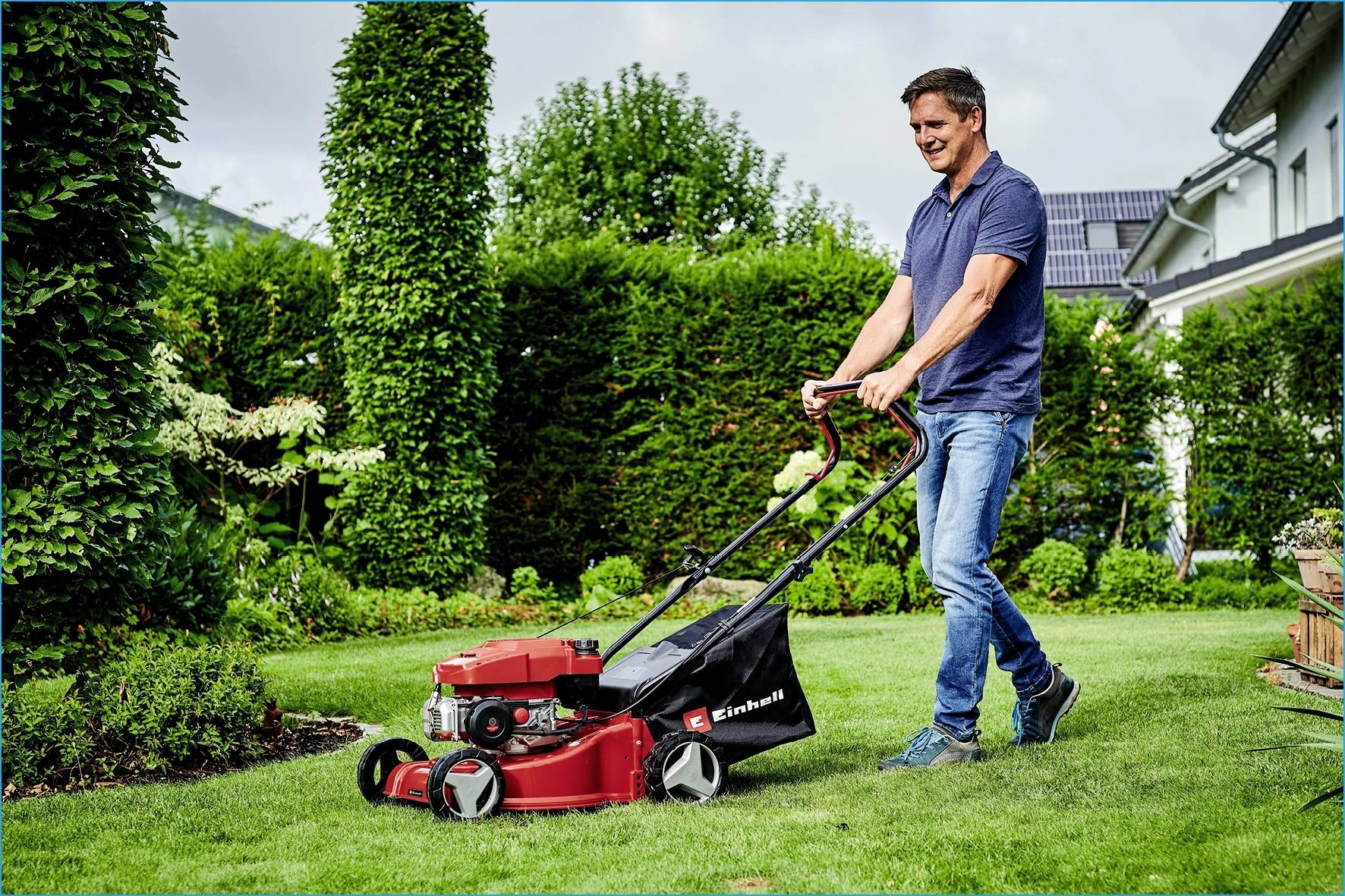 Ein Mann mäht Rasen mit einem roten Rasenmäher im Garten. Im Hintergrund sind Büsche, Bäume und ein Haus zu sehen.