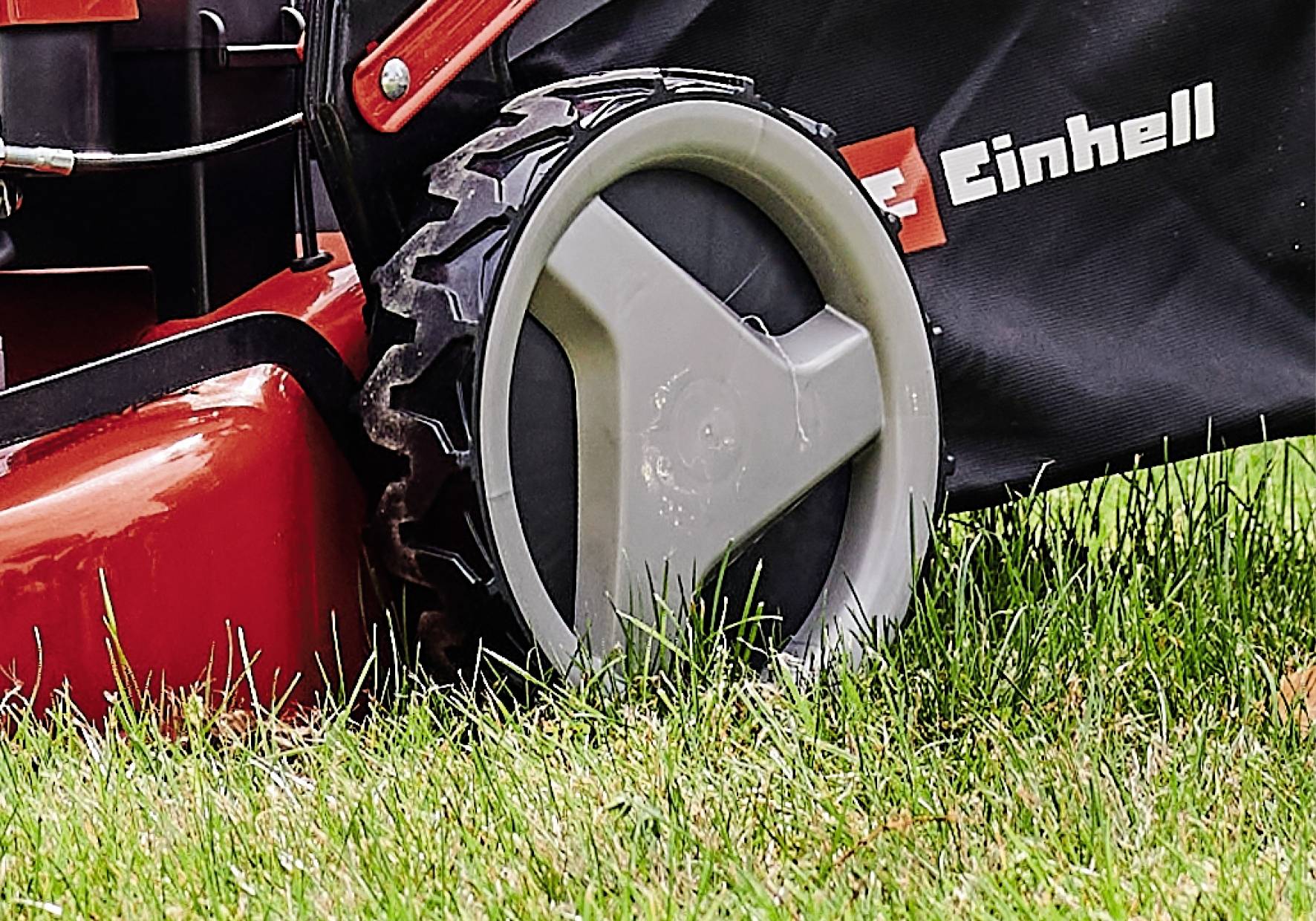 Rasenmäher auf grünem Gras mit Fokus auf das Rad und einen Teil des Gehäuses. Die Marke 'Einhell' ist auf der Abdeckung sichtbar.