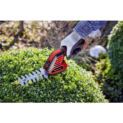 Eine Person schneidet mit einer roten elektrischen Heckenschere einen grünen Busch. Die Hand trägt einen Gartenhandschuh aus Stoff.