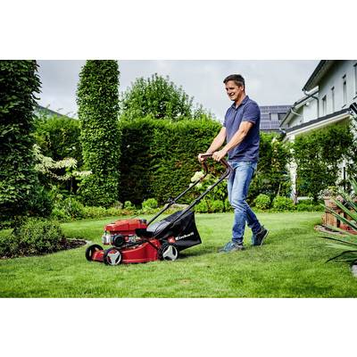 Ein Mann mäht den Rasen in einem Garten mit einem roten Rasenmäher. Im Hintergrund sind Büsche, Bäume und ein Haus sichtbar.