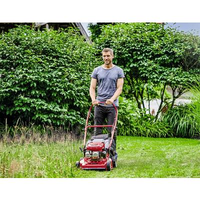 Mann mäht Rasen mit einem roten Rasenmäher in einem grünen Garten. Hintergrund: Büsche und Bäume.