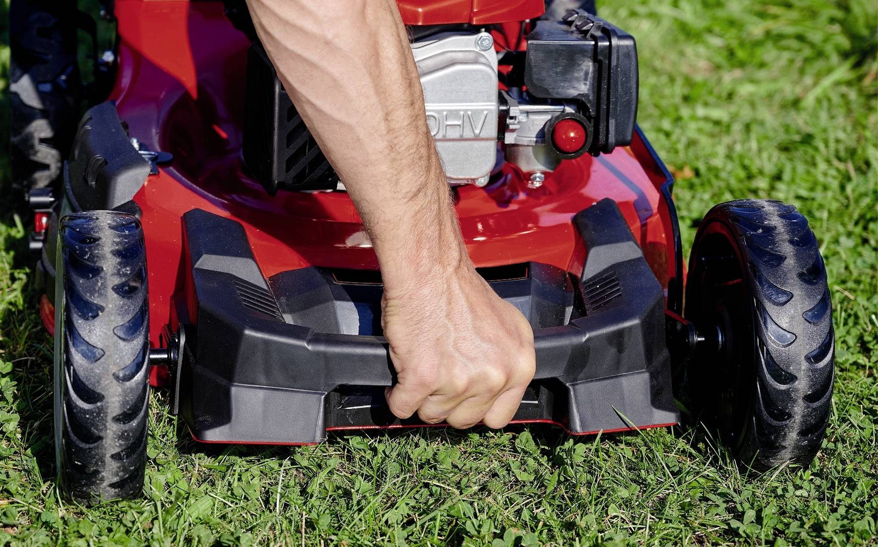 Eine Person hält einen Rasenmähergriff, während sie ihn über einen grünen Rasen schiebt. Der Mäher ist rot mit großen schwarzen Rädern.