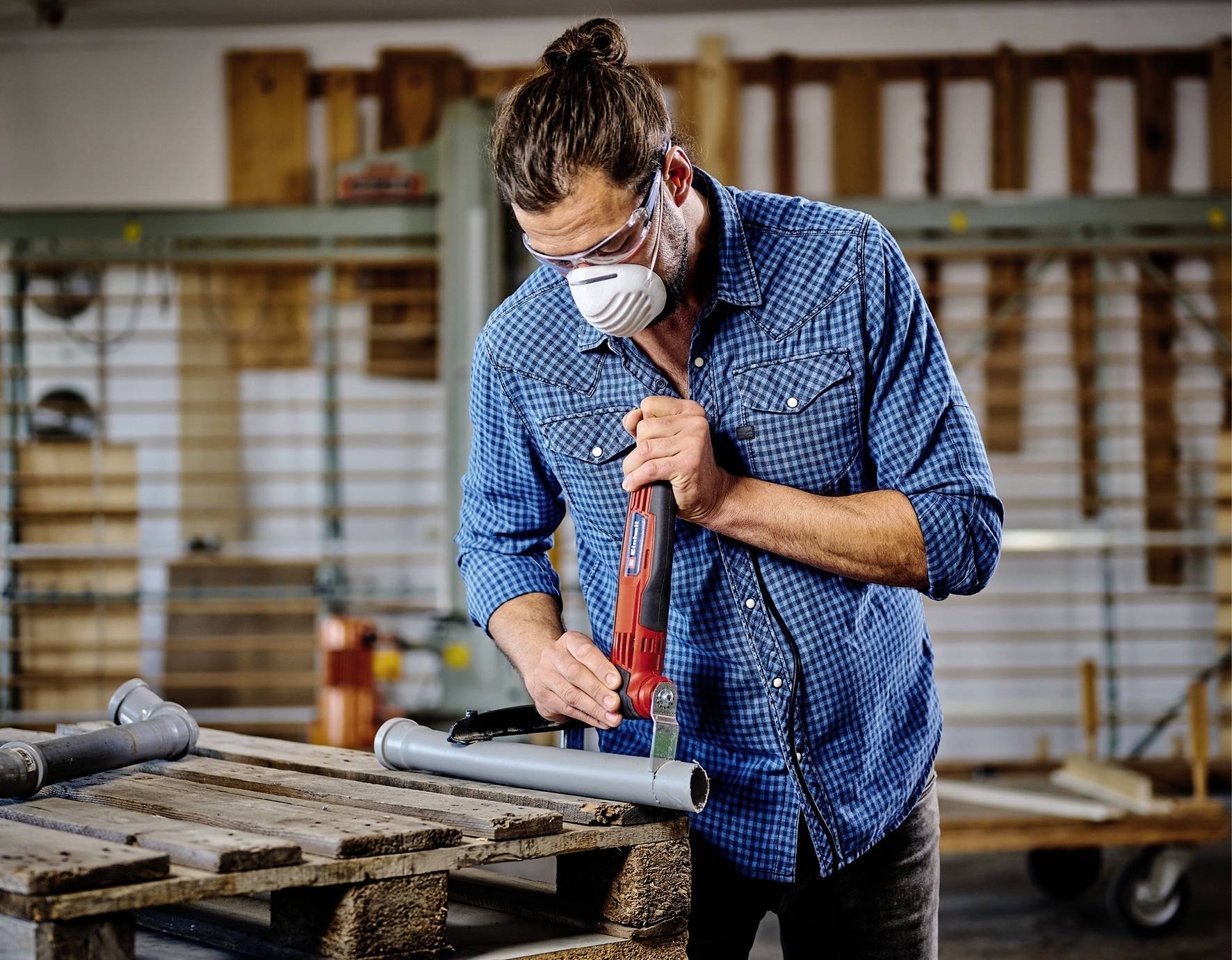 Ein Mann in Schutzbrille und Mundschutz bearbeitet ein Metallrohr mit einer Elektrosäge in einer Werkstatt.