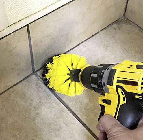 Eine Hand hält einen gelben Akkuschrauber mit einer Bürstenaufsatz, der Fliesenfugen reinigt. Im Hintergrund sind Fliesen zu sehen.