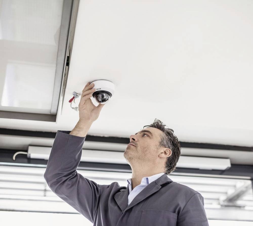 Ein Mann in Arbeitskleidung untersucht oder installiert eine Überwachungskamera an der Decke in einem modernen Gebäude.