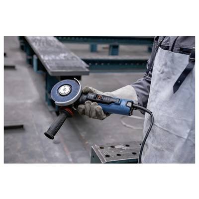 Eine Person mit Handschuhen und Schürze hält einen blauen Winkelschleifer und steht auf einem industriellen Boden mit Metallträgern im Hintergrund.