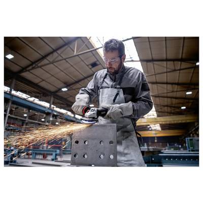 Ein Mann in Schutzausrüstung schleift ein Metallstück mit einem Elektrowerkzeug in einer Industriewerkstatt, während Funken sprühen und Maschinen im Hintergrund zu sehen sind.