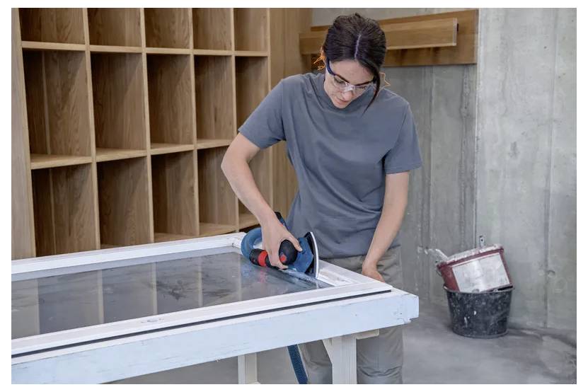 Eine Person benutzt ein Elektrowerkzeug, um die Kanten eines Holzrahmens auf einem Tisch in einer Werkstatt zu glätten. Im Hintergrund befinden sich Regale und ein Eimer.