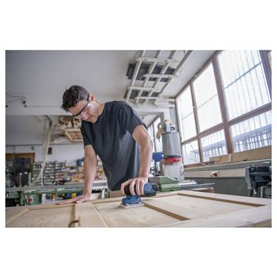 Eine Person mit Sicherheitsbrille schleift eine Holzoberfläche in einer Werkstatt. Verschiedene Werkzeuge und Ausrüstungsgegenstände sind im Hintergrund zu sehen.