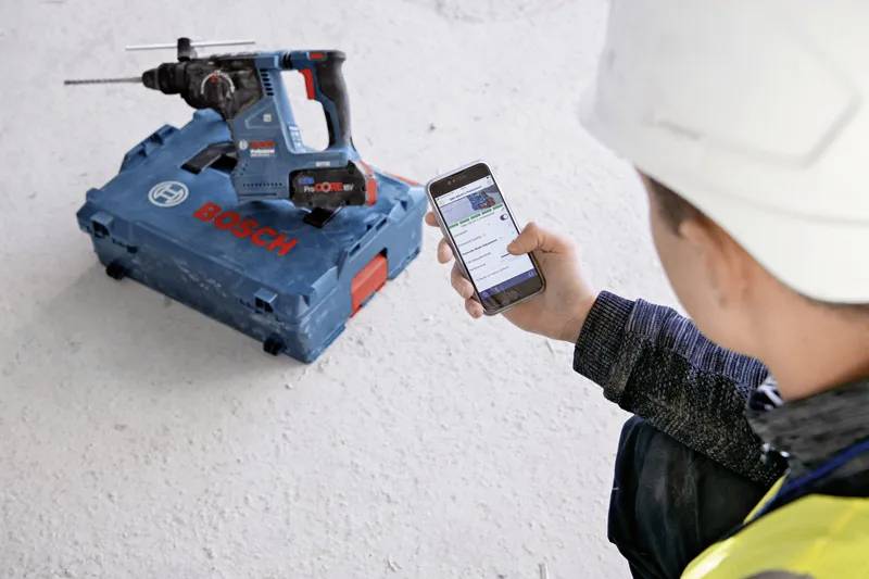 Eine Person in Arbeitskleidung der Baubranche untersucht eine mobile App, die Details eines Bosch-Werkzeugs anzeigt, das in der Nähe auf dem Boden liegt.