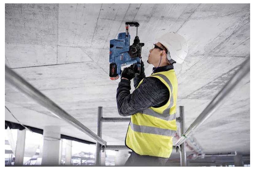 Ein Bauarbeiter in Schutzausrüstung benutzt ein Elektrowerkzeug, um eine Betondecke auf einem Gerüst in einem unfertigen Gebäude zu bohren.