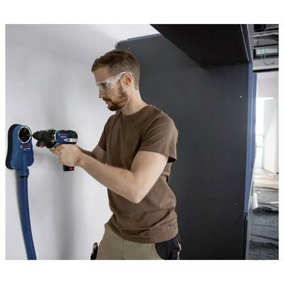 Eine Person, die eine Schutzbrille trägt, verwendet einen Elektroschrauber mit Staubabsaugung an einer weißen Wand.