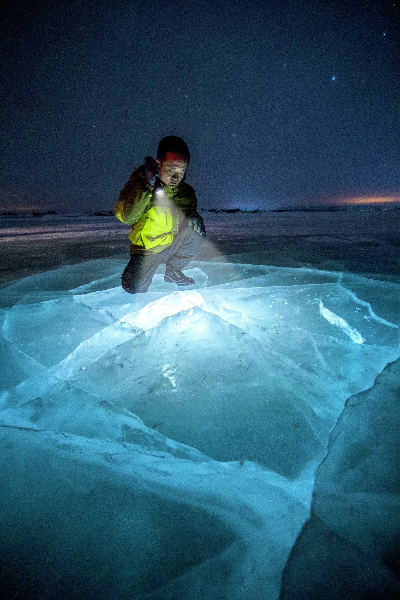 Eine Person in Winterkleidung kniet auf einer leuchtenden Eisfläche bei Nacht und beleuchtet sie mit einer Taschenlampe für eine Untersuchung.