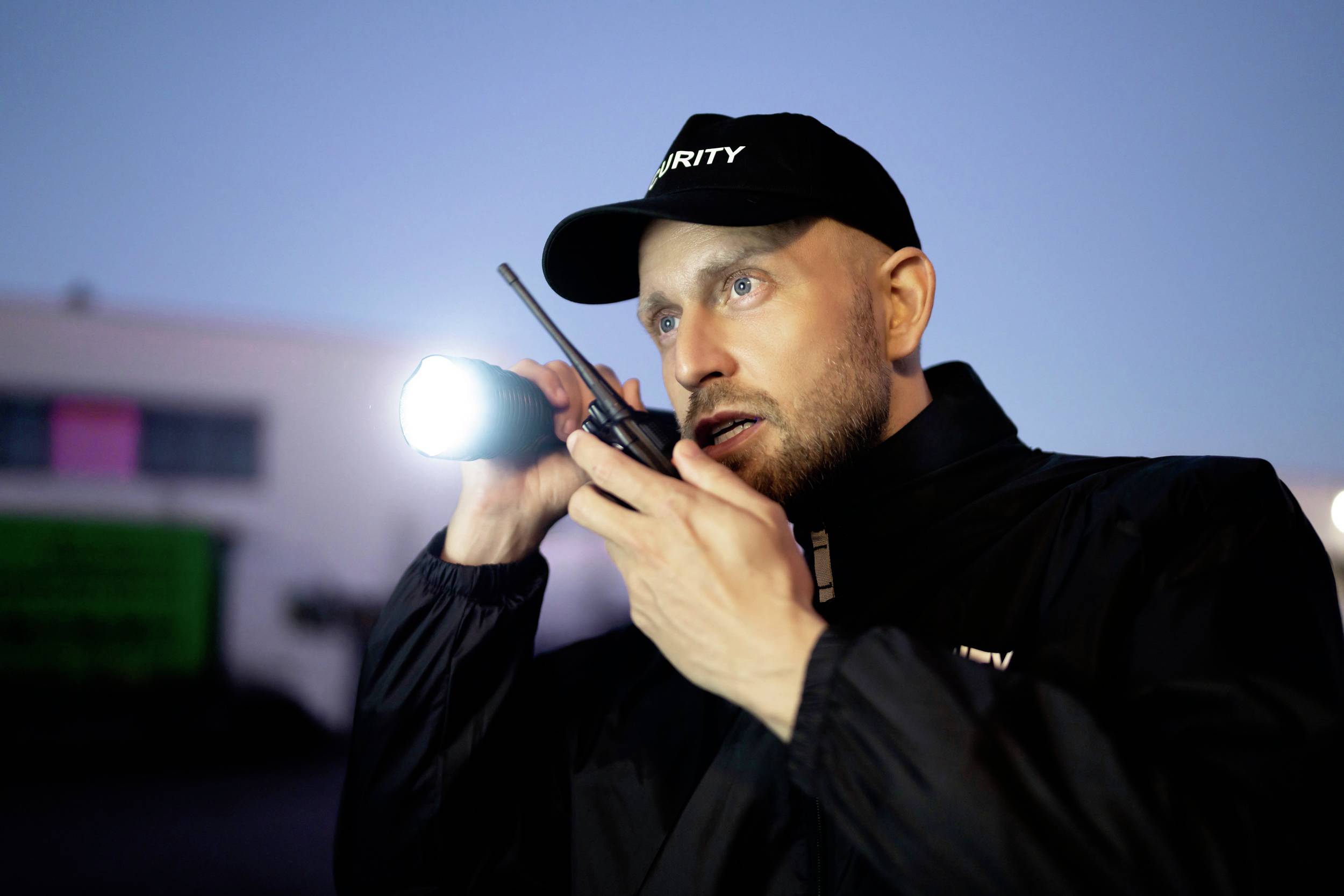 Ein Wachmann in Uniform steht nachts im Freien, hält eine Taschenlampe und spricht in ein Funkgerät.