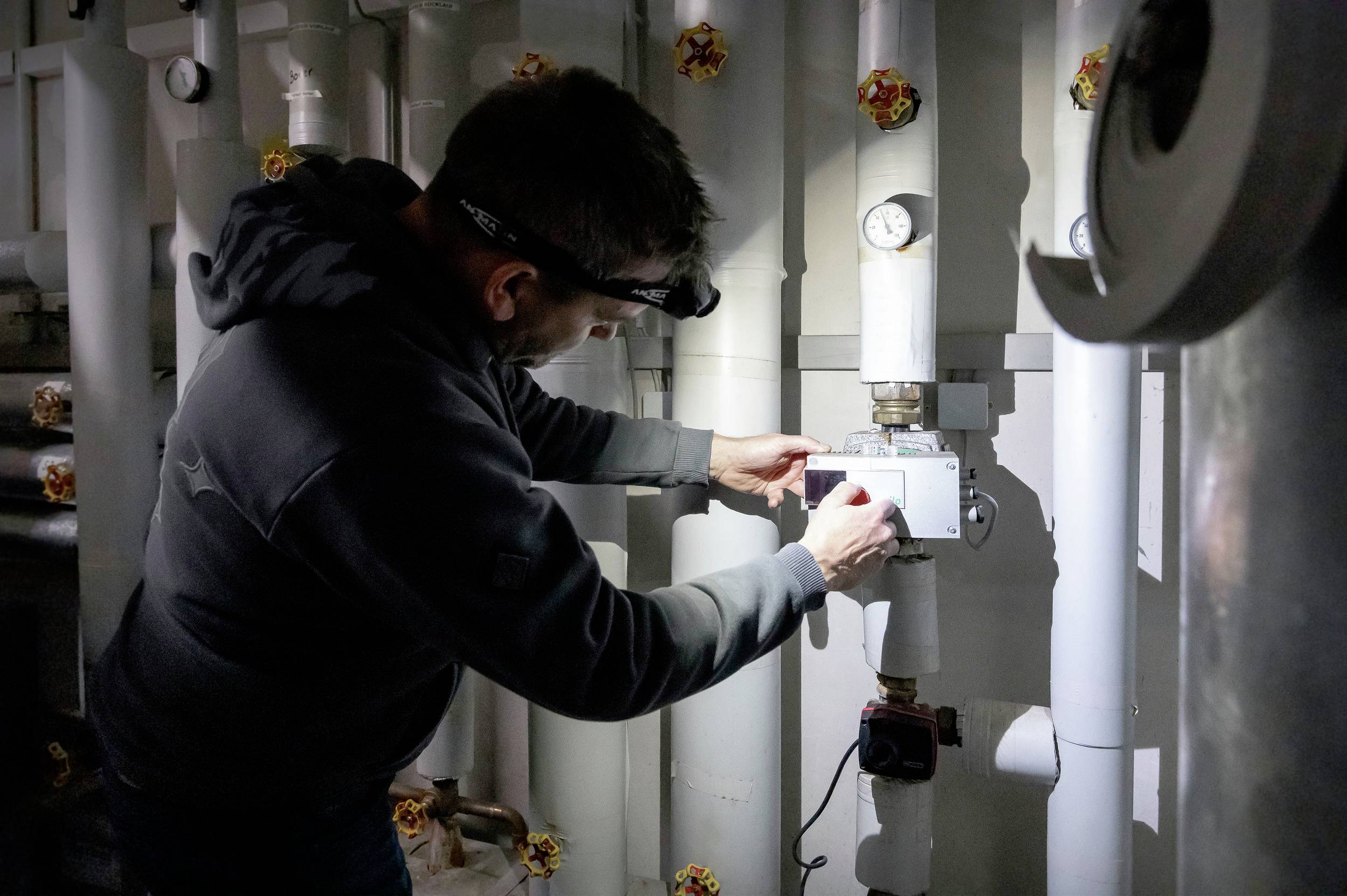 Ein Techniker in dunkler Kleidung arbeitet mit einer Stirnlampe in einem Maschinenraum an einer Steuereinheit an einem Rohrleitungssystem.