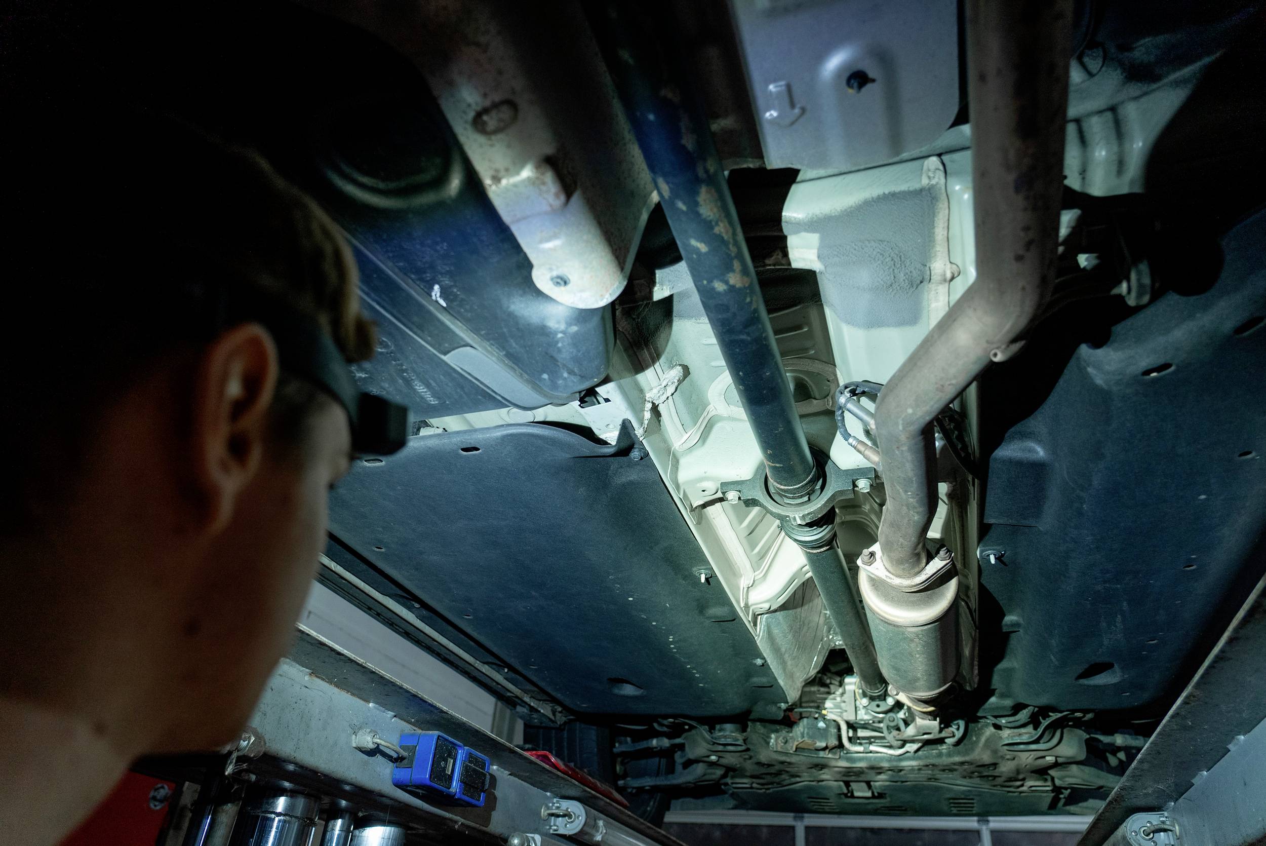 Eine Person betrachtet das Fahrgestell eines Autos in einer Werkstatt von unten. Verschiedene Autoteile und Rohrleitungen sind sichtbar.