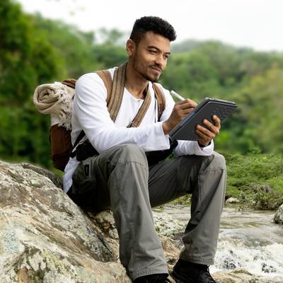 Ein Mann sitzt auf einem Felsen in der Natur, hält ein Notizbuch oder Tablet in der Hand und wirkt konzentriert.