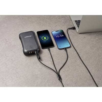 Vier Geräte sind auf einer Betonoberfläche verbunden: Eine Powerbank, zwei aufladende Smartphones und ein Laptop.